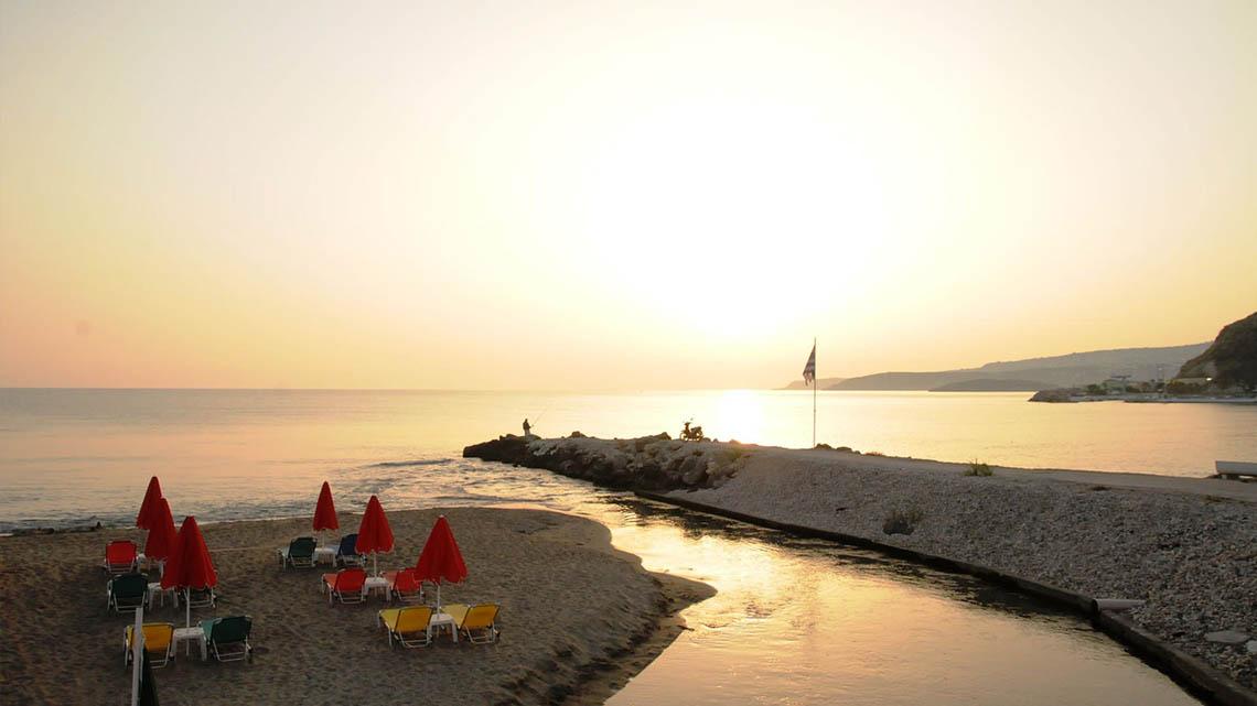 Sonnenuntergang am Strand des Kalyves Beach Hotels mit Liegestühlen und roten Sonnenschirmen.