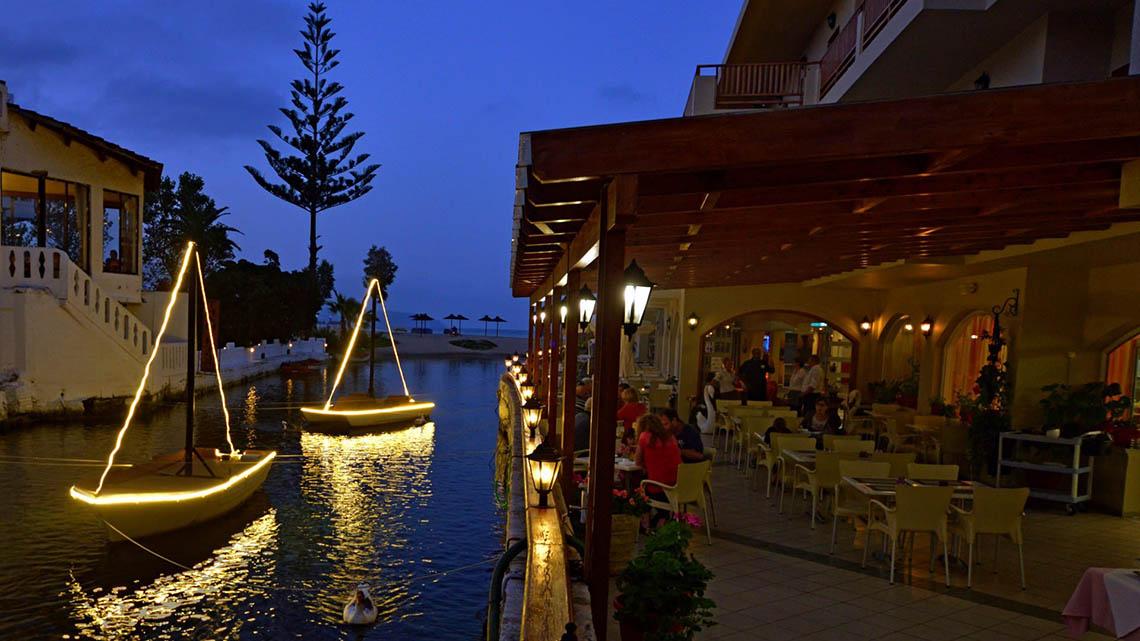 Abendansicht des Kalyves Beach Hotels mit beleuchteten Booten und Restaurant am Wasser.