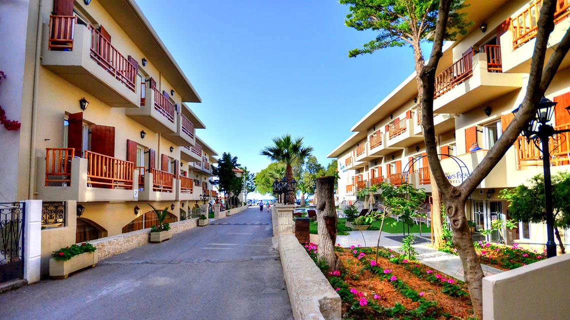 Die Allee des Kalyves Beach Hotels mit gepflegten Gärten und hohen Gebäuden.