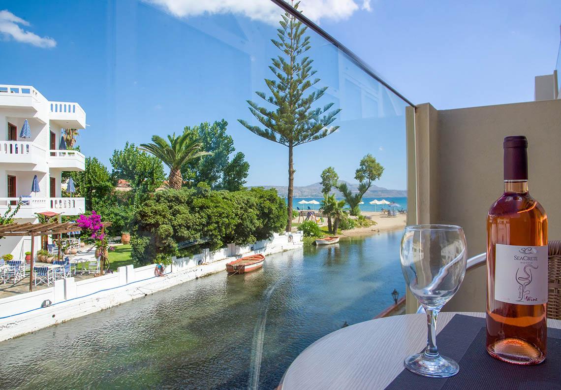 Aussicht von Kalyves Beach Hotel auf Strand und Kanal mit einem Glas und einer Flasche Wein auf dem Tisch.