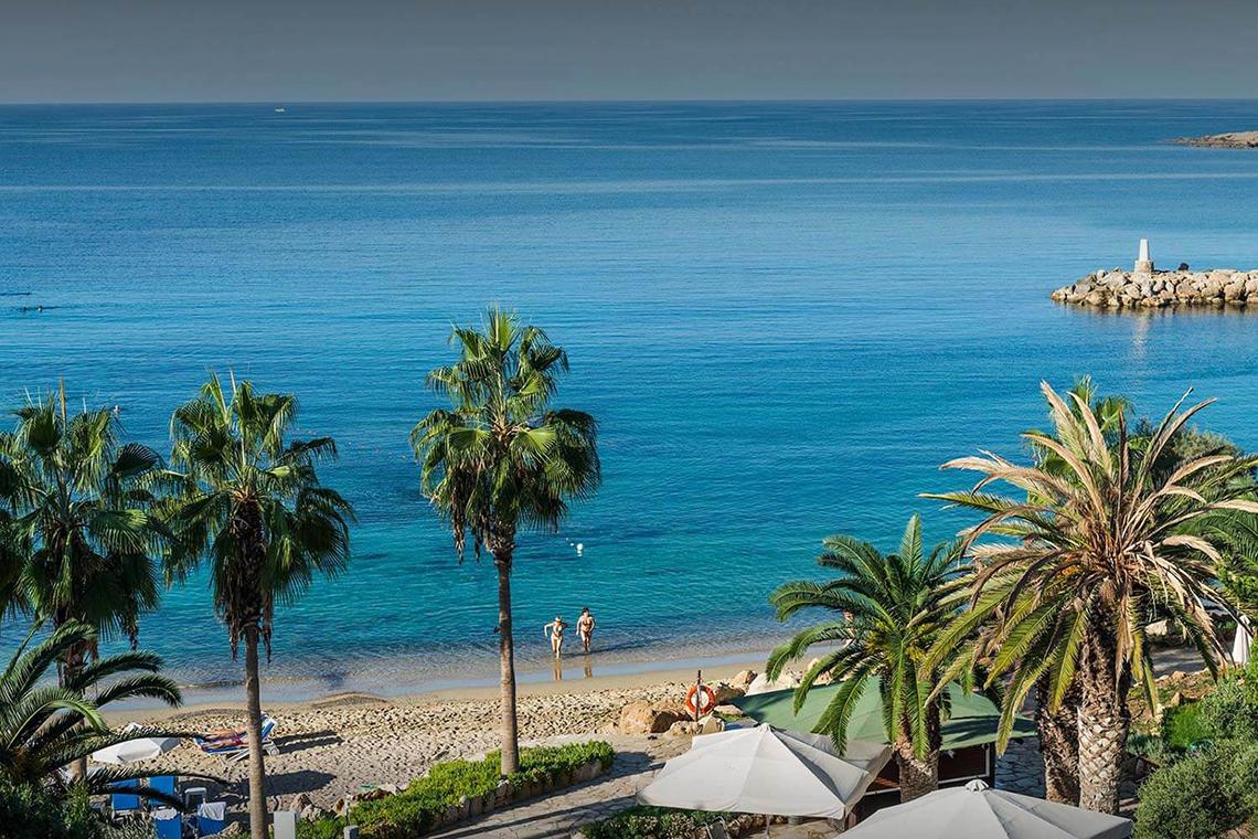 Ausblick auf den Strand des Coral Beach Hotel & Resorts mit zwei Personen, die im Wasser spazieren gehen und vielen Palmen
