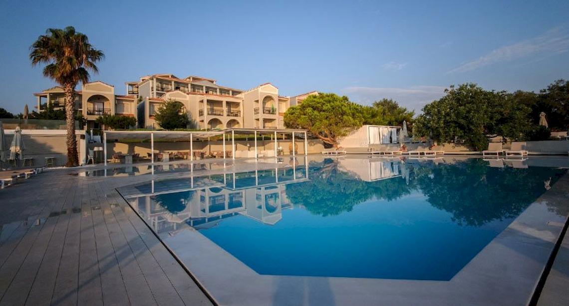 Der Swimmingpool des Bay Hotel & Suites mit Sonnenliegen und Bäumen im Hintergrund.