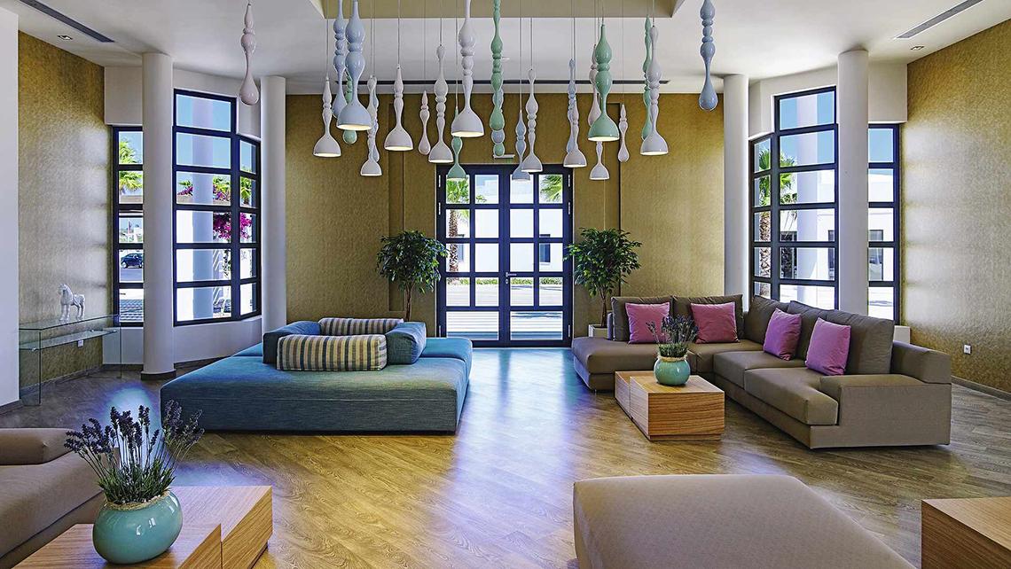 Lobby des Club Plimmiri Hotels mit modernen Möbeln, Holzboden und großen Fenstern.