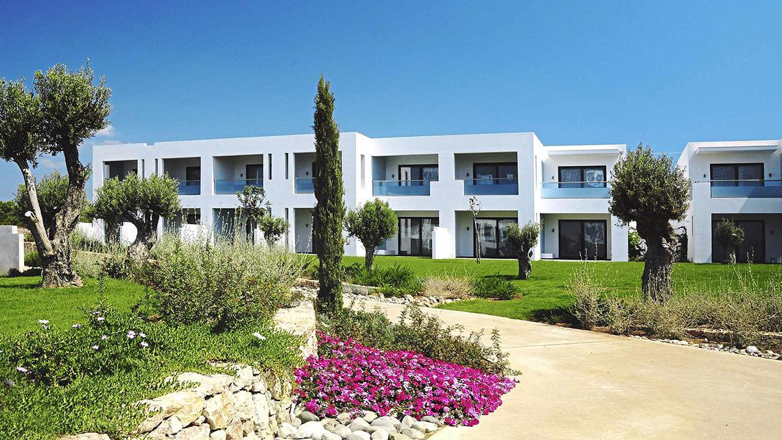 Außenansicht des weißen Club Plimmiri Hotels mit gepflegtem Rasen und Bäumen davor.
