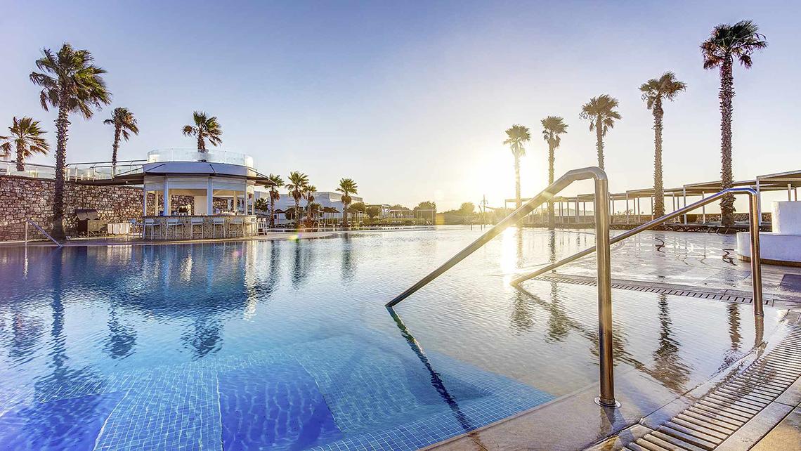 Sonnenuntergang am Pool des Club Plimmiri Hotels mit Palmen und einer Bar im Hintergrund.