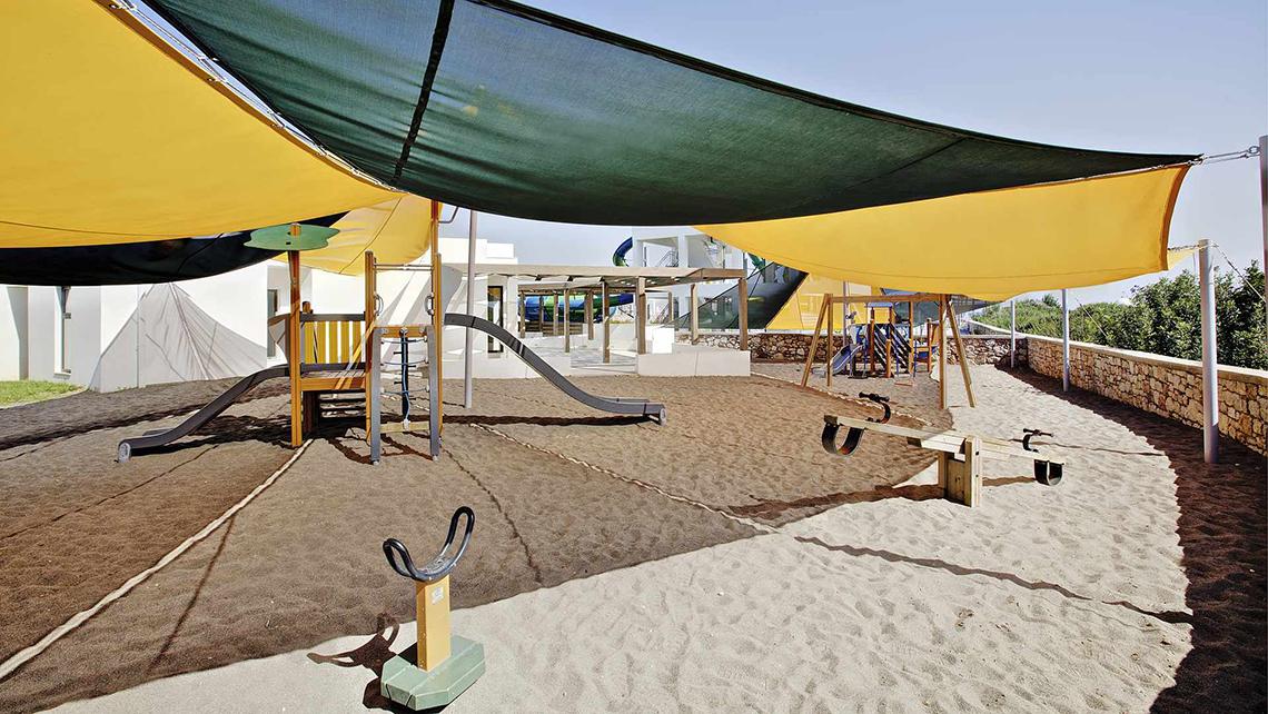 Kinderspielplatz im Club Plimmiri Hotel mit Sand, Rutschen und Schaukeln unter einem Sonnensegel.
