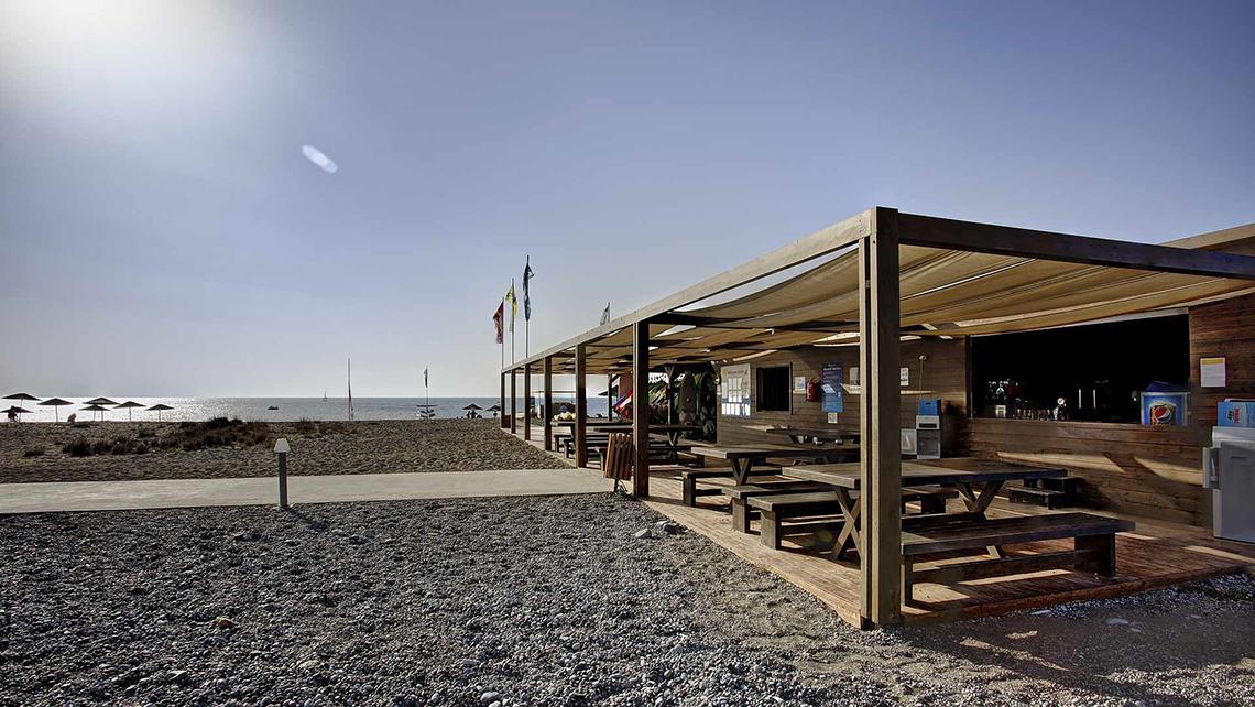 Außenbereich des Club Plimmiri Hotels mit Holztischen und Bänken unter einem Sonnendach am Strand.