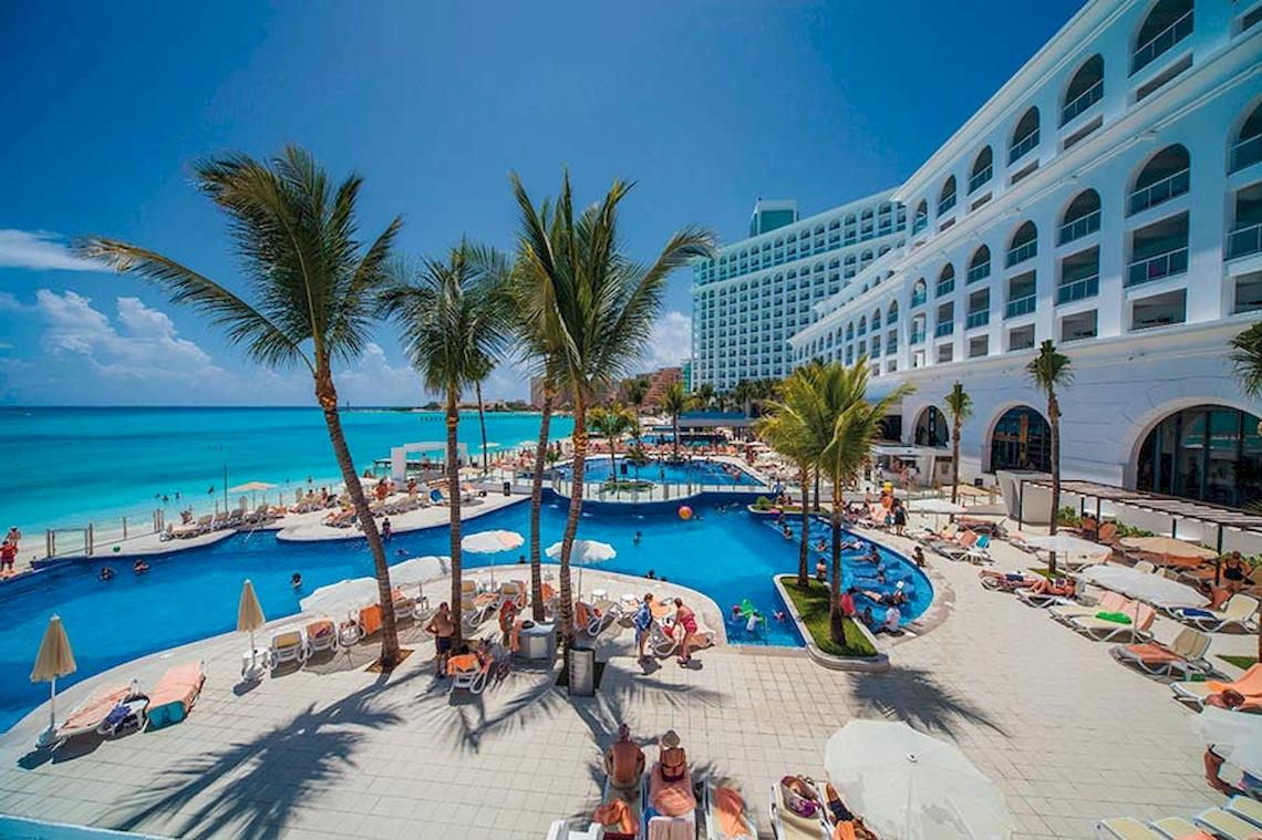 Einer der Pools des Riu Cancun Hotels mit vielen Gästen, Sonnenliegen und Palmen.