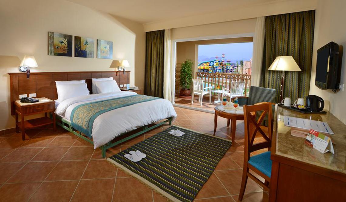 Charmillion Club Aqua Park Hotelzimmer mit Bett, Teppich, Schreibtisch und Balkon mit Wasserrutsche im Hintergrund.