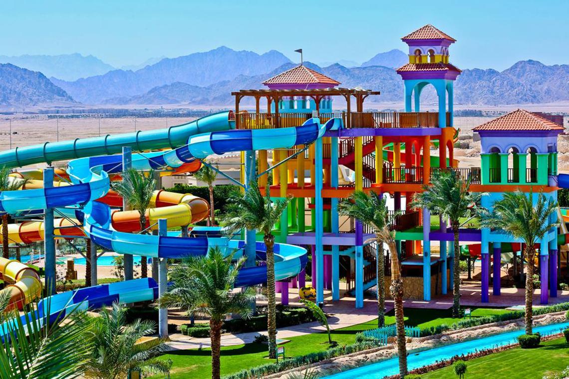Wasserrutschen im Charmillion Club Aqua Park mit bunten Gebäuden und Bergen im Hintergrund.