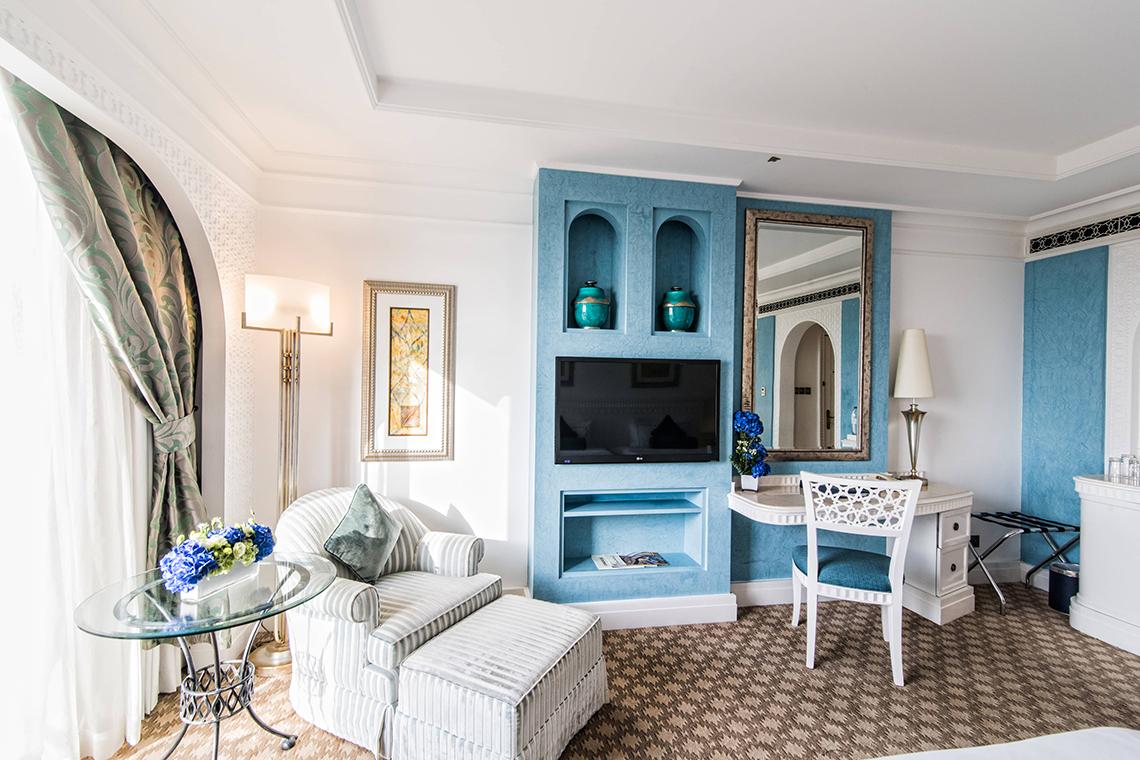 Innenansicht eines blau dekorierten Zimmers im Habtoor Grand Resort & Spa mit Fernseher, Schreibtisch und Stühlen.