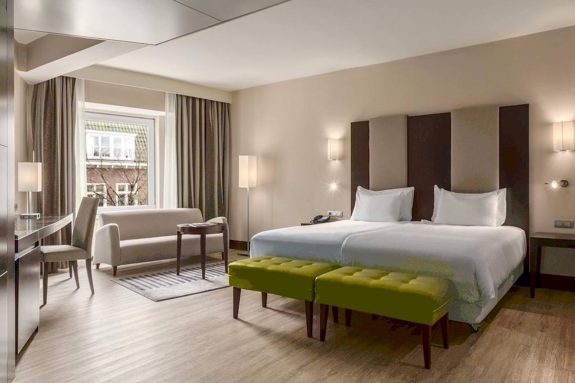 Modernes Zimmer im NH Amsterdam Centre Hotel mit zwei Betten, grünen Hockern und einem Fenster.