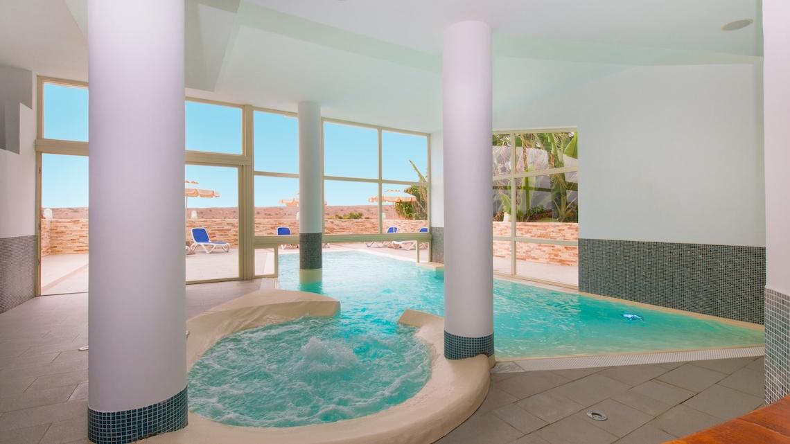 Innenpool im VOI Praia De Chaves Resort mit Sonnenliegen und Fenstern, die zum Außenbereich führen.
