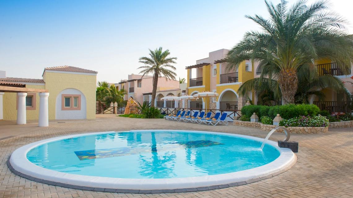Schwimmbecken im VOI Praia De Chaves Resort mit blauen Liegestühlen und gelben Gebäuden im Hintergrund.