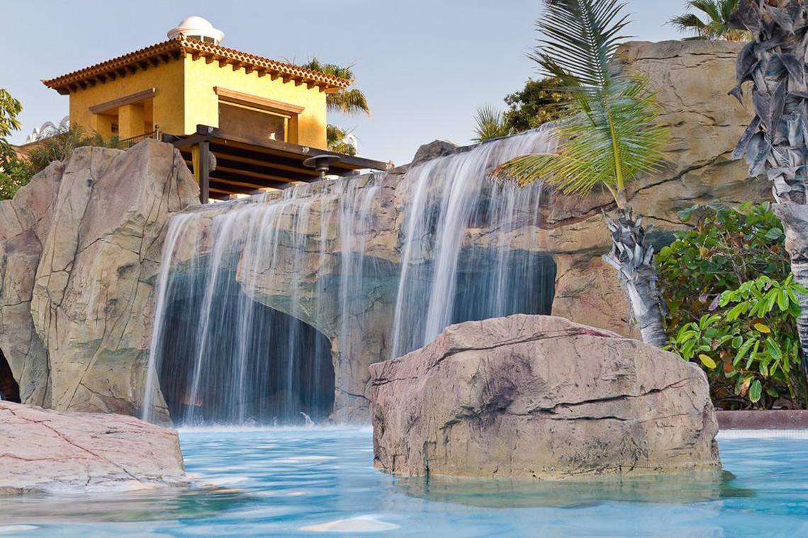 Wasserfall und Poolbereich der Europa Villa Cortes mit einer gelben Villa im Hintergrund.