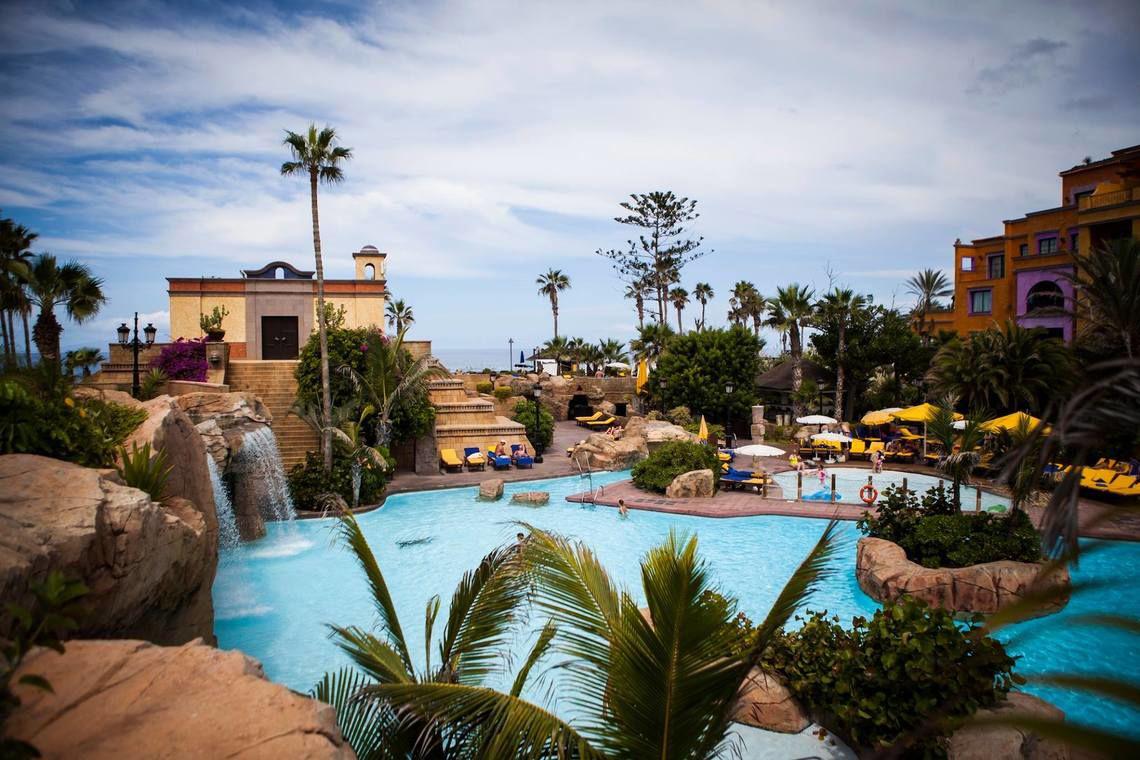 Außenpoolbereich des Europe Villa Cortes Hotels mit Wasserfällen, Sonnenliegen und gelben Sonnenschirmen.