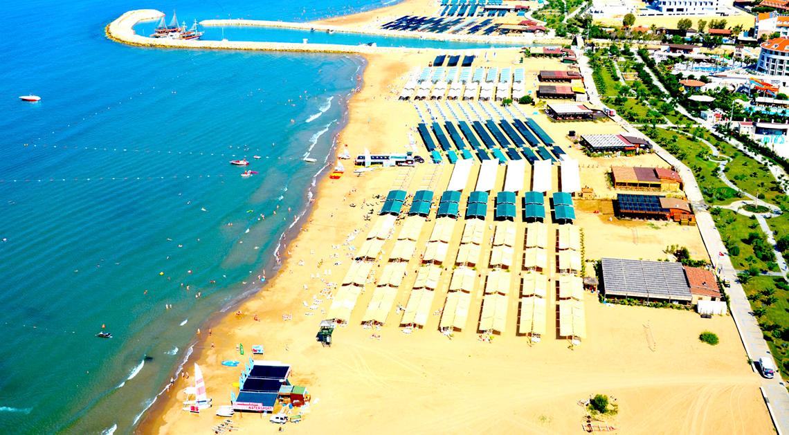 Luftaufnahme des Port Side Resorts, das an einem Strand liegt, mit vielen Sonnenschirmen und Sonnenliegen.