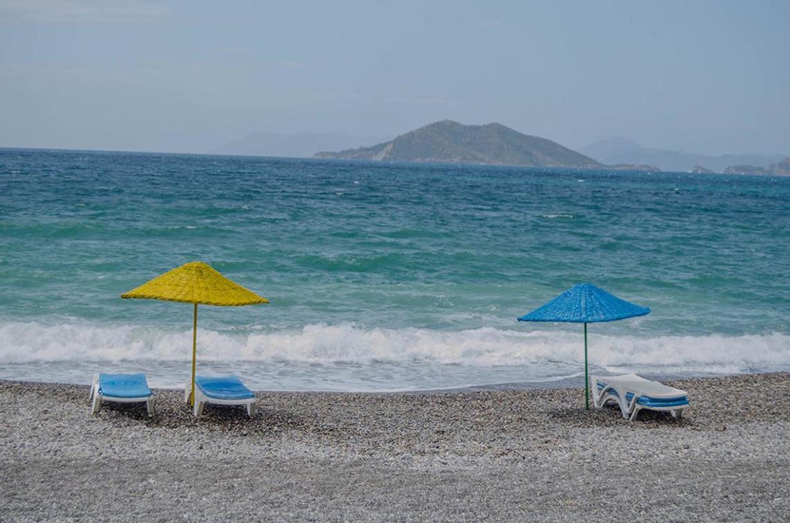 Strandszene am Malhun Hotel mit zwei Sonnenliegen und Sonnenschirmen, vor einem ruhigen Meer und einer kleinen Insel im Hintergrund.