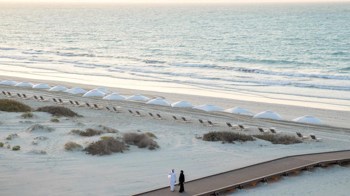 Strand des Jumeirah at Saadiyat Island Resorts mit weißen Sonnenschirmen und zwei Personen auf dem Gehweg.