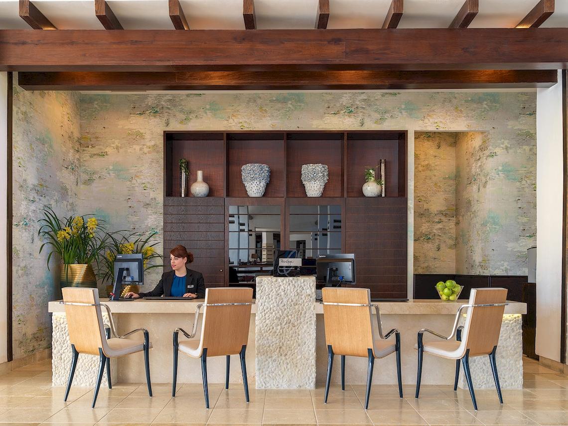 Empfangsbereich des H10 Estepona Palace Hotels mit Rezeptionistin, Vasen und Pflanzen im Hintergrund.