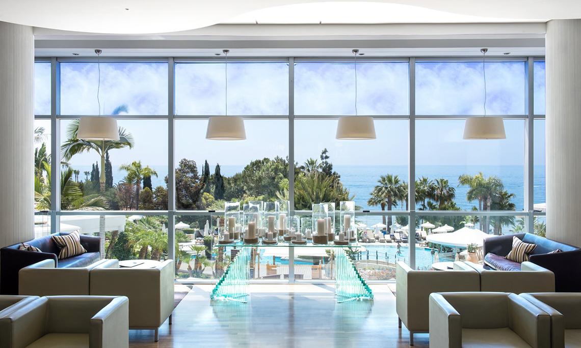 Lobby des Mediterranean Beach Hotels mit Blick auf den Ozean, große Fenster, moderne Möbel und eine Glastisch mit Kerzen.