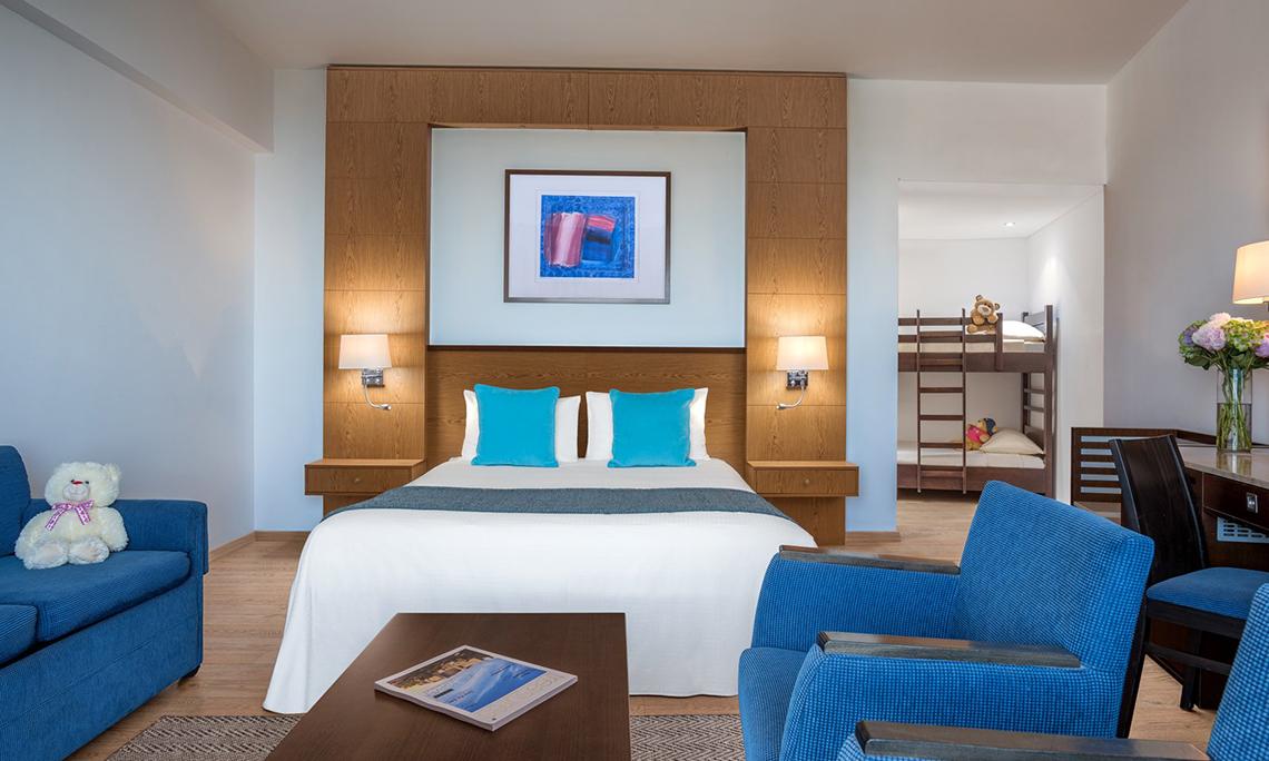 Innenansicht eines Zimmers im Mediterranean Beach Hotel in Zypern mit einem Bett, zwei blauen Kissen, einem Sofa, einem Tisch, einem Teddybär und einem Bild an der Wand.