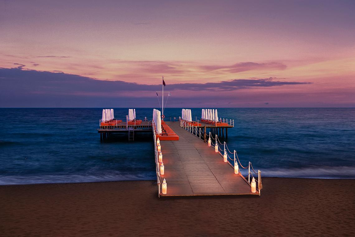 Aska Lara Resort & Spa Pier am Meer bei Sonnenuntergang mit roten Liegen und Laternen