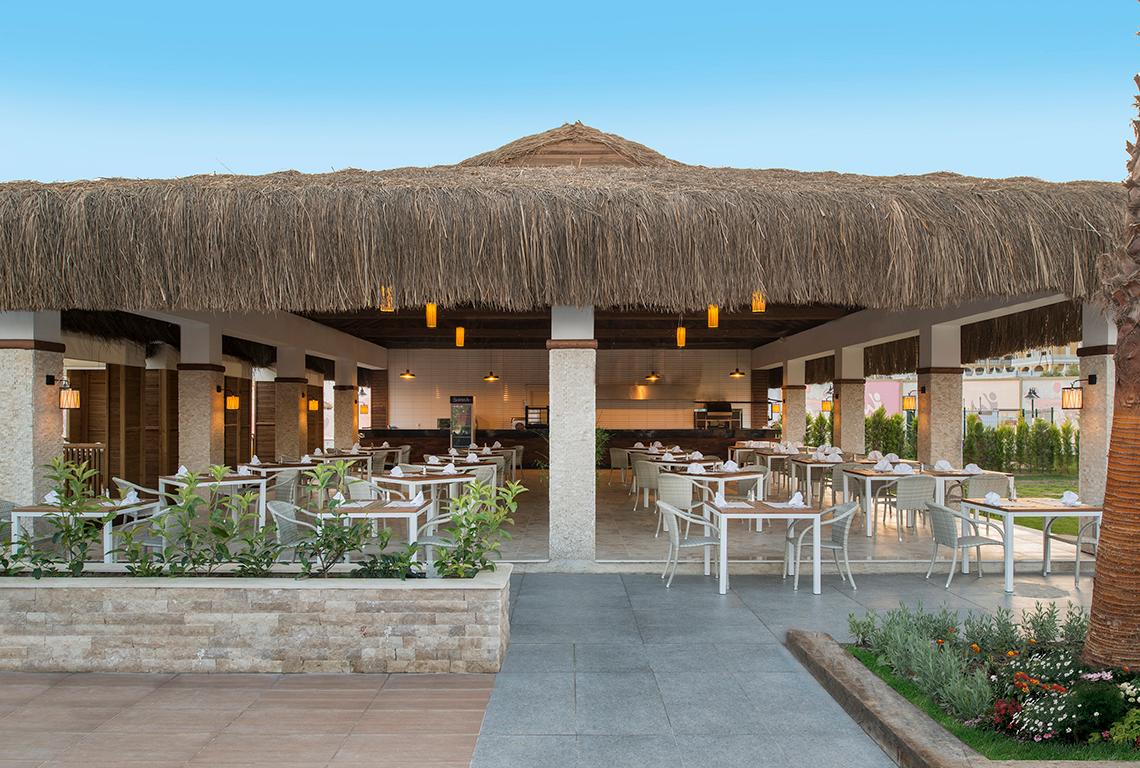 Außenbereich des Aska Lara Resort & Spa mit rustikalem Strohdach und runden Tischen und Stühlen.