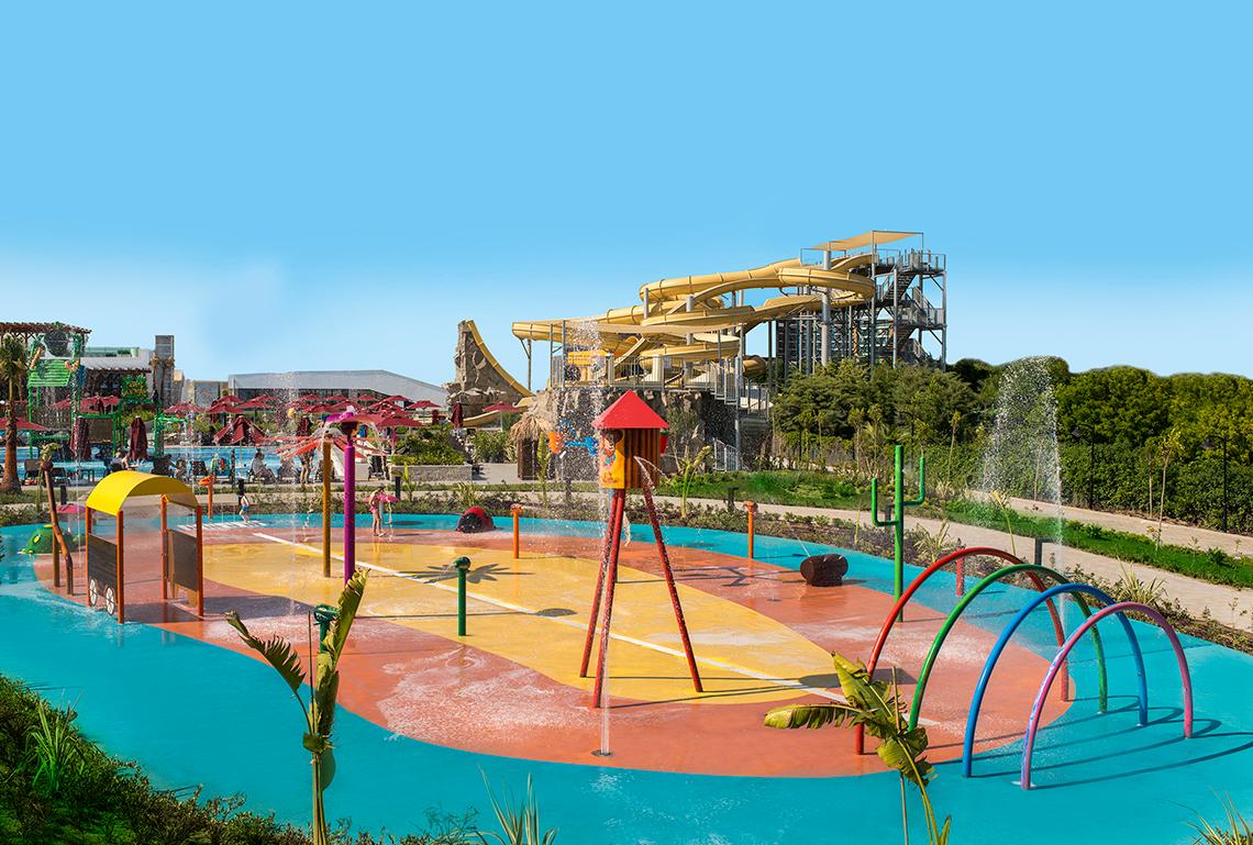 Wasserpark des Aska Lara Resort & Spa mit bunten Spielgeräten und einer großen Wasserrutsche im Hintergrund.