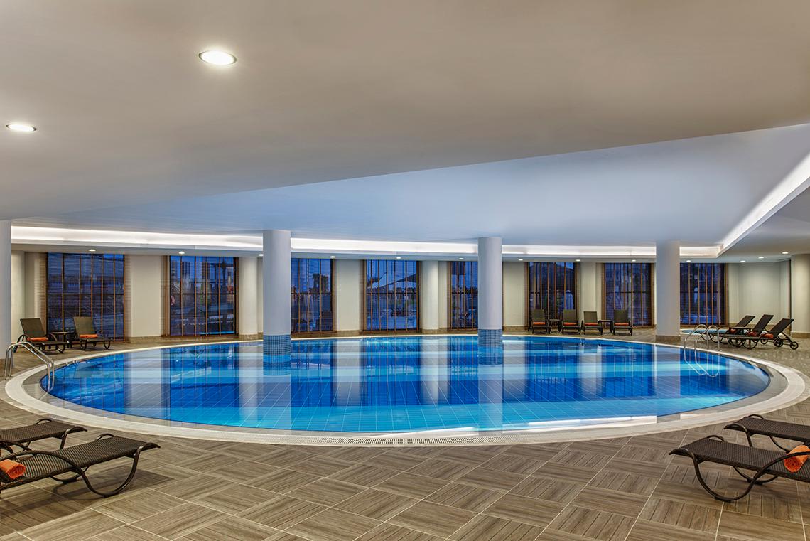 Innenpool des Aska Lara Resort & Spa mit Liegestühlen und Panoramafenstern