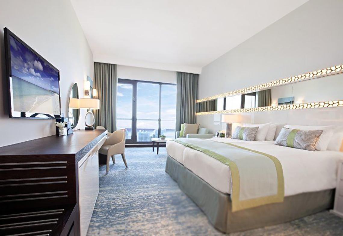 Innenansicht eines Zimmers im JA Ocean View Hotel mit großem Bett, Fernseher und Meerblick.