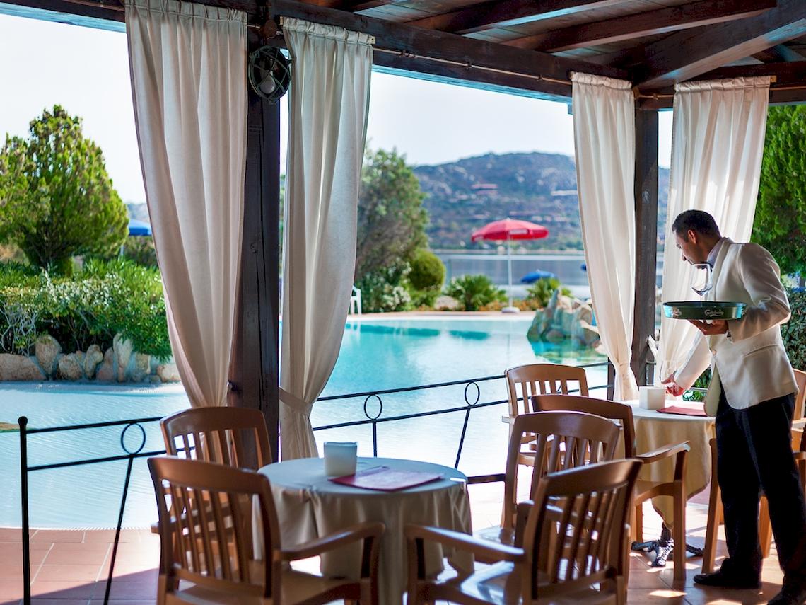 Ein Kellner im Colonna Hotel Du Golf serviert ein Tablett mit einem Glas Wein auf der Terrasse mit Poolblick.