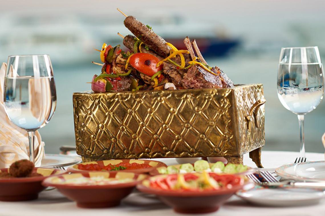 Luxuriöses Essen im Continental Hotel Hurghada mit gegrilltem Fleisch und Gemüse in einer goldenen Schale, serviert mit Wein und Beilagen.