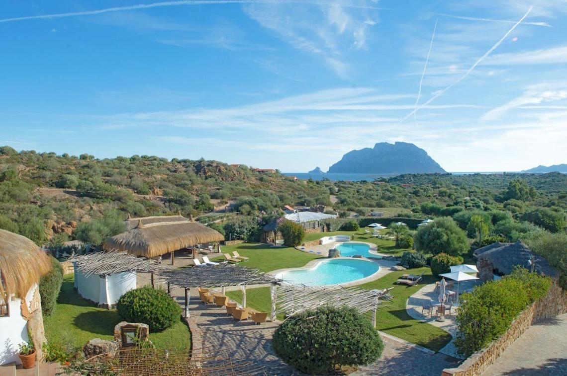 Luftaufnahme des Hotel Ollastu Resorts mit Swimmingpool, Sonnenliegen und Bergpanorama.