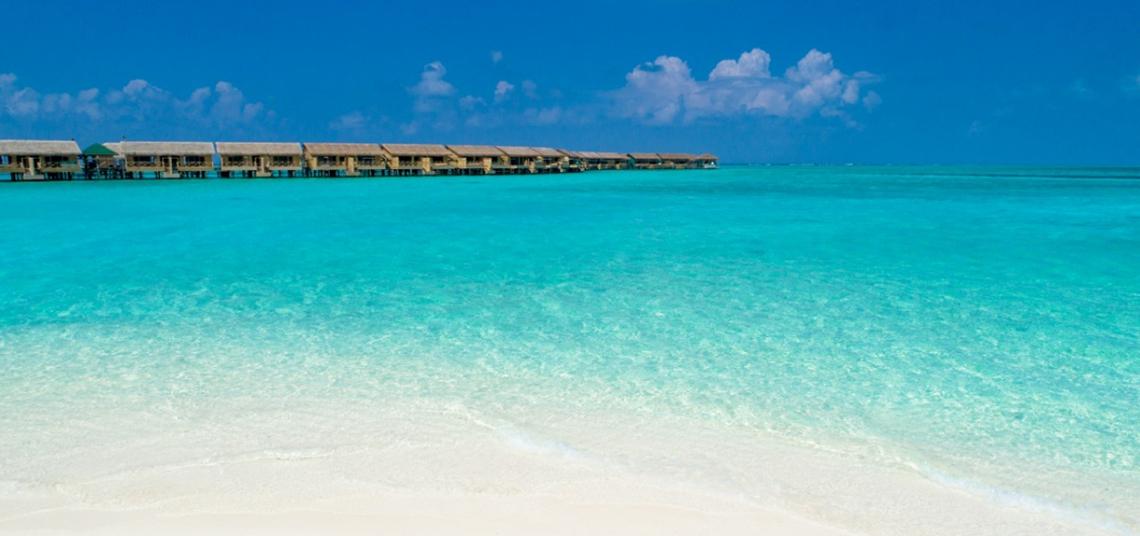 Strandszene mit kristallklarem Wasser und weißen Sand, mit Hotelhütten im Hintergrund