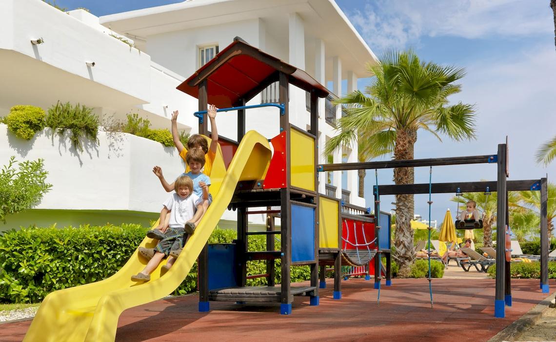 Kinder spielen auf einem bunten Spielplatz im Freien, im Hintergrund steht das H10 Estepona Palace Hotel.