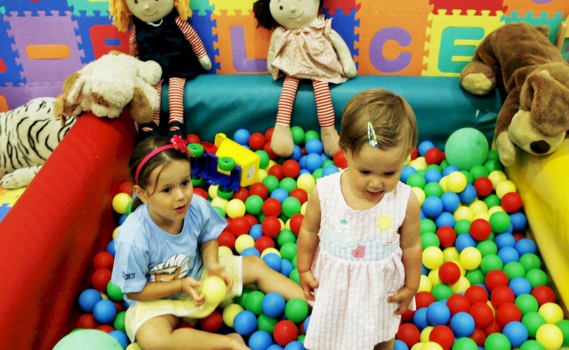 Zwei Mädchen spielen in einem Ballbad mit bunten Bällen, während Puppen und Spielzeuge im Hintergrund stehen.