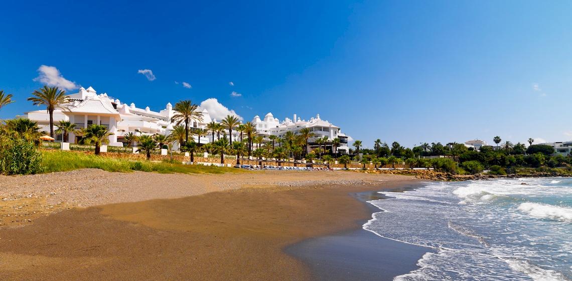 Außenansicht des H10 Estepona Palace Hotels mit Blick auf eine lange, helle Sandstrand und das Meer.