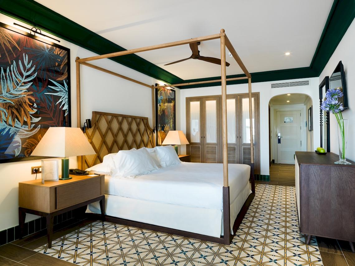 Schlafzimmer im H10 Estepona Palace mit Himmelbett, weißen Laken, zwei Nachttischen und tropischen Wandgemälden.