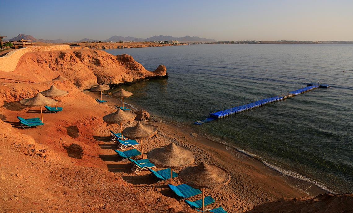 Strand des Sharm Club Beach Resorts mit Sonnenliegen, Sonnenschirmen und einer Brücke im Wasser.
