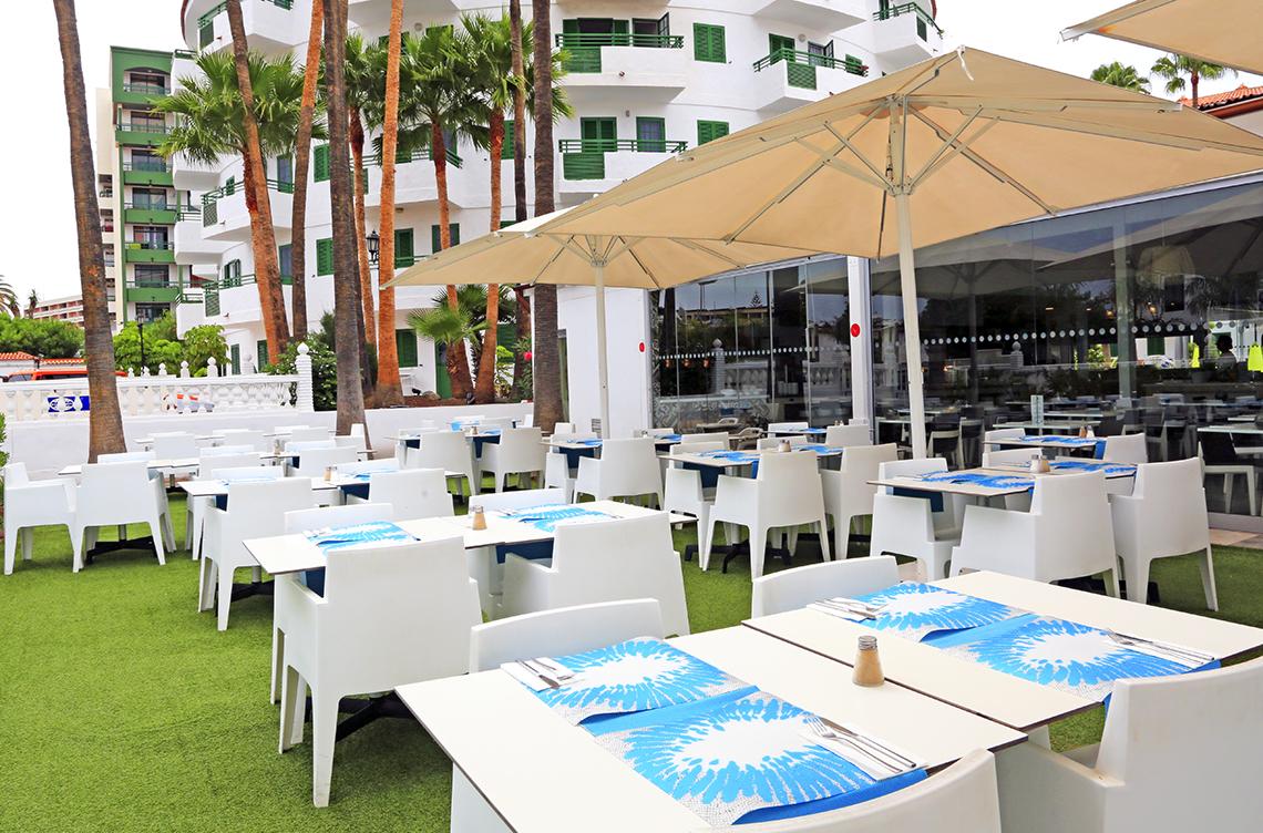 Außenbereich des Restaurants des Hotels Servatur Playa Bonita mit weißen Tischen und Stühlen, blauem Geschirrtuch und großen Sonnenschirmen