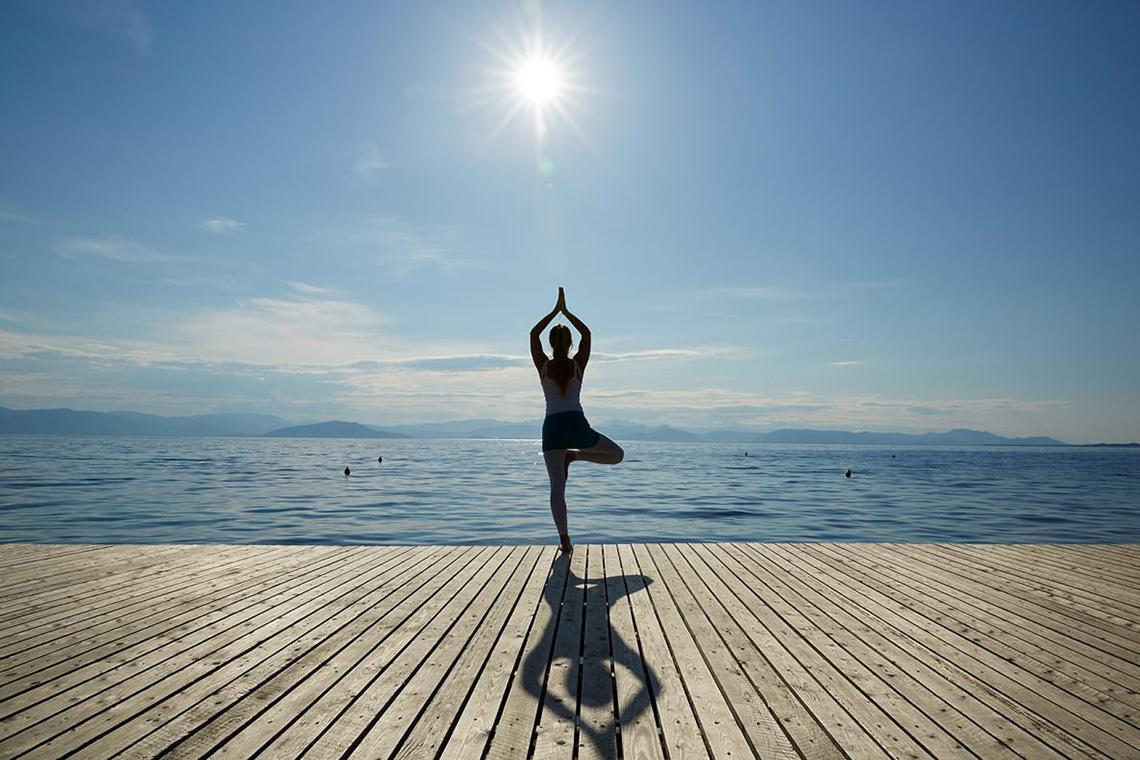 Eine Person praktiziert Yoga in einer Balance-Pose auf einer Holzplattform am Meer.