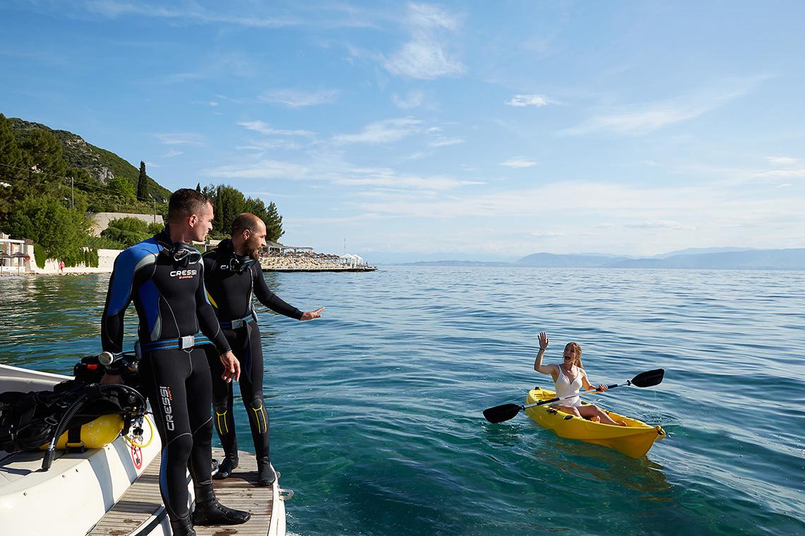 Zwei Taucher stehen am Dock, während eine Frau in einem gelben Kajak auf dem Wasser paddelt.