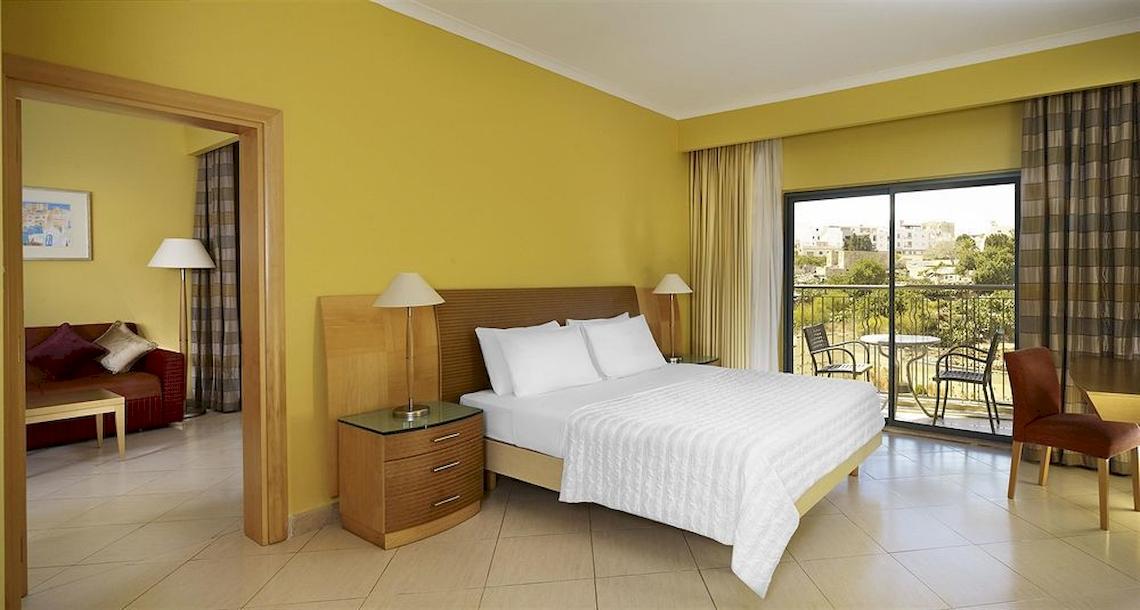 Ein helles Hotelzimmer mit gelben Wänden, einem Bett, einem Balkon, und Möbeln.