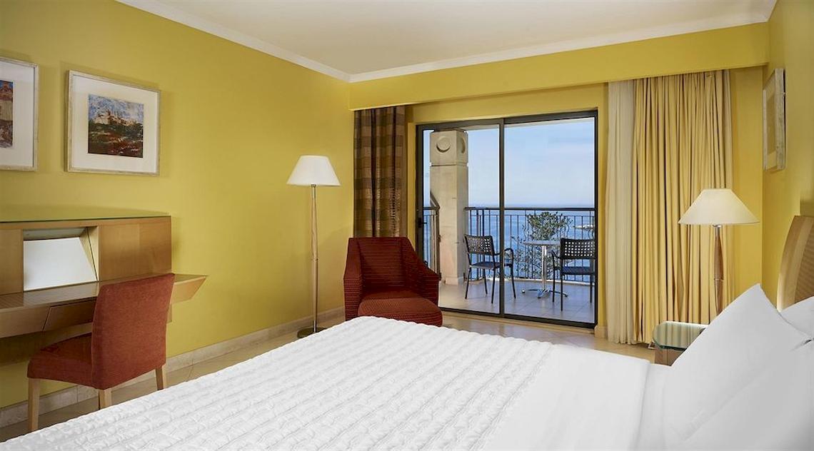 Ein modernes Hotelzimmer mit gelben Wänden, einem Bett, Schreibtisch, Stühlen und einer Terrasse mit Meerblick.