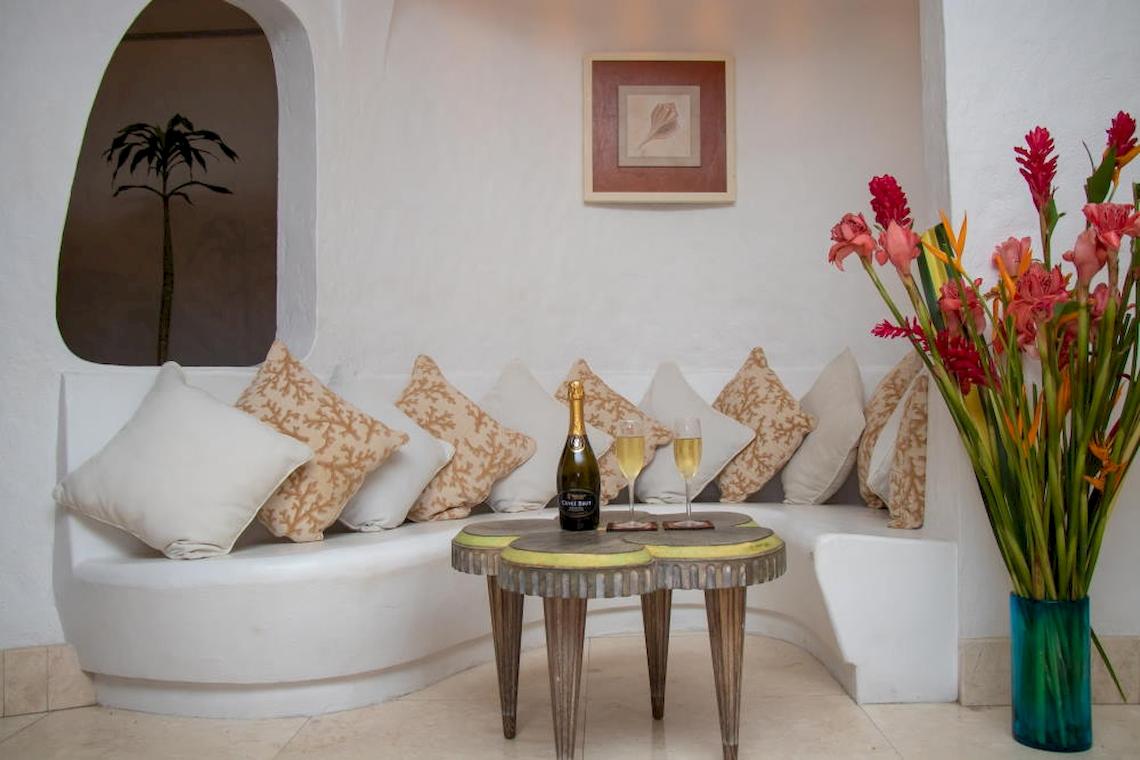 Loungebereich im Village du Pecheur Hotel mit weißer Sitzbank, Kissen, Flasche Champagner und zwei Gläsern auf einem Tisch neben bunten Blumen in einem blauen Vasen.