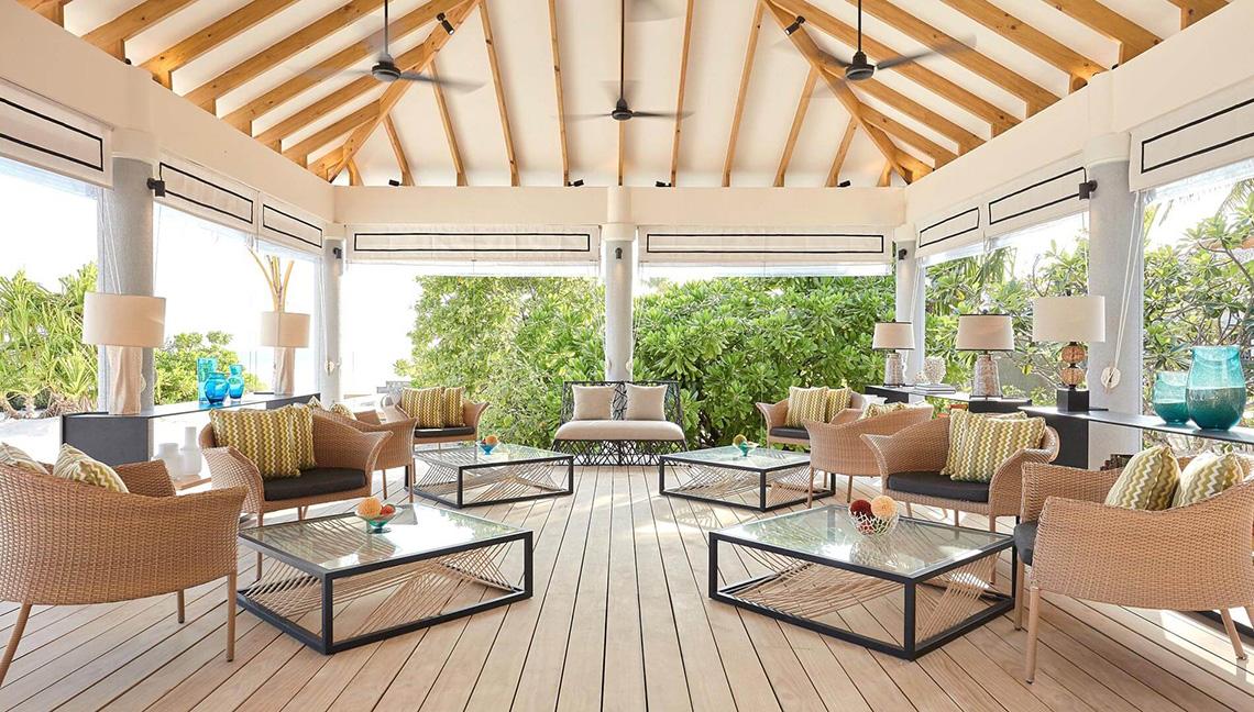 Lounge-Bereich des Amari Havodda Maldives mit Holzboden, modernen Möbeln und großen Fenstern, die einen Blick auf die Außenlandschaft gewähren.