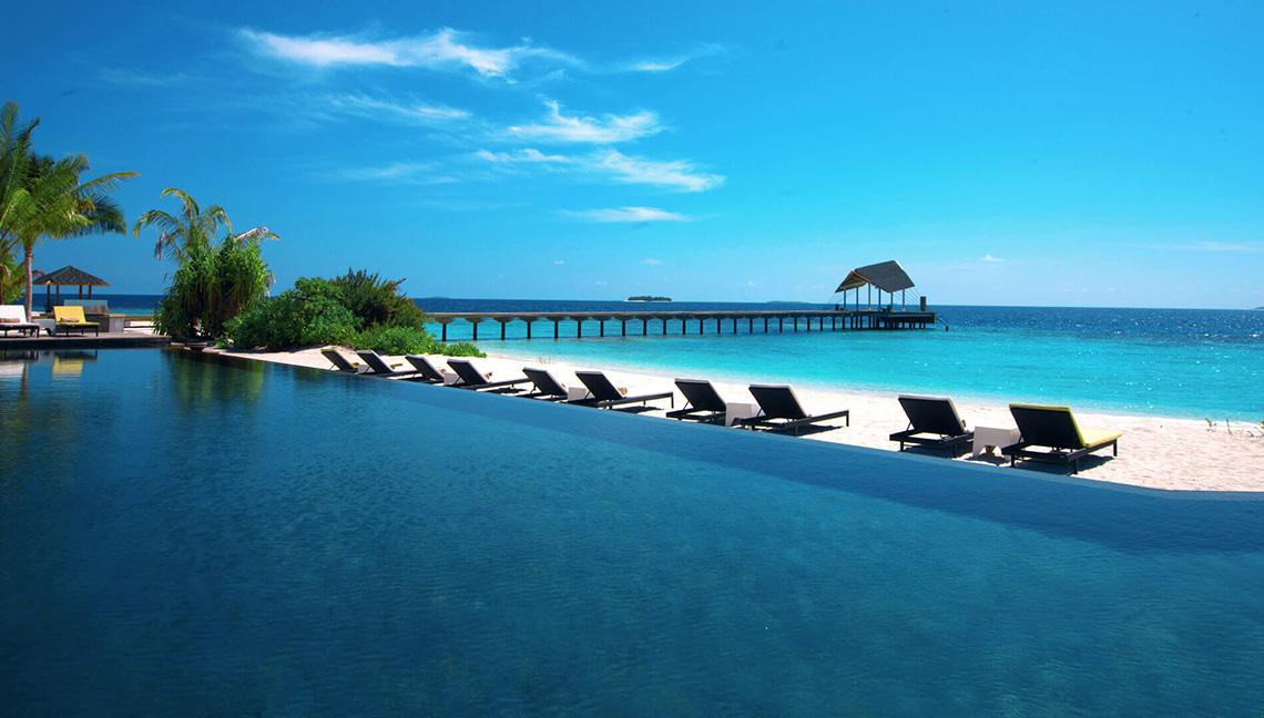Ein großer Pool am Strand des Amari Havodda Maldives mit Sonnenliegen und einer Pier im Hintergrund.