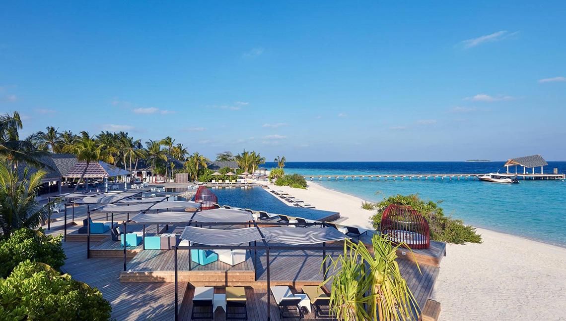 Amari Havodda Maldives Strand mit mehreren Liegebereichen und einem Pier im Hintergrund.