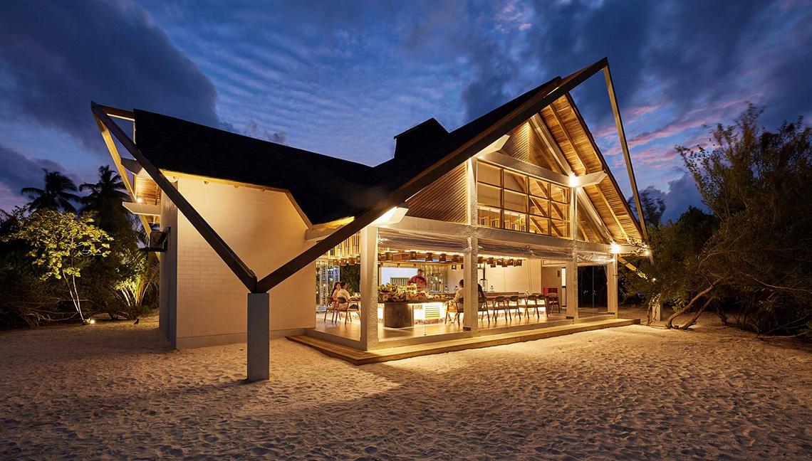 Außenansicht des Amari Havodda Maldives Hotel bei Nacht mit beleuchteter Fassade und Gästen im Restaurantbereich.